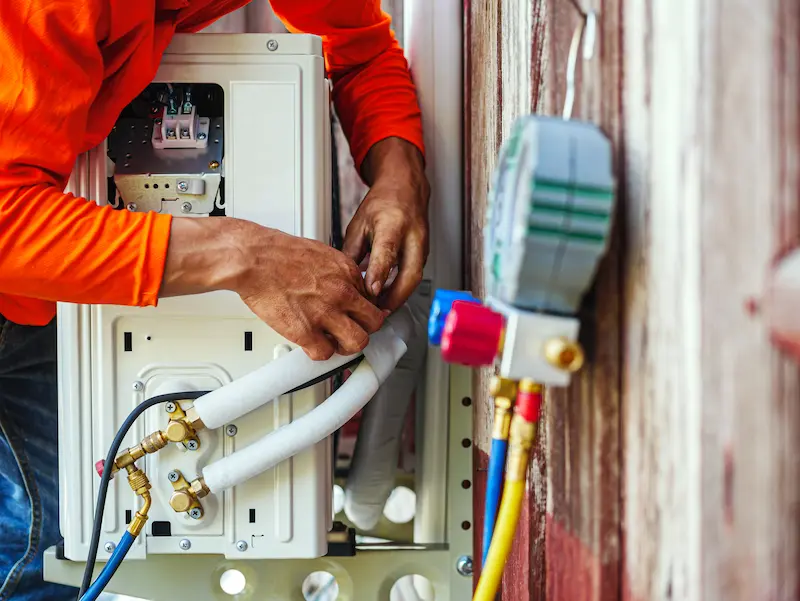 An HVAC technician in an orange shirt servicing an outdoor heat pump split system unit, connecting refrigerant lines with a manifold gauge in Brownsville.