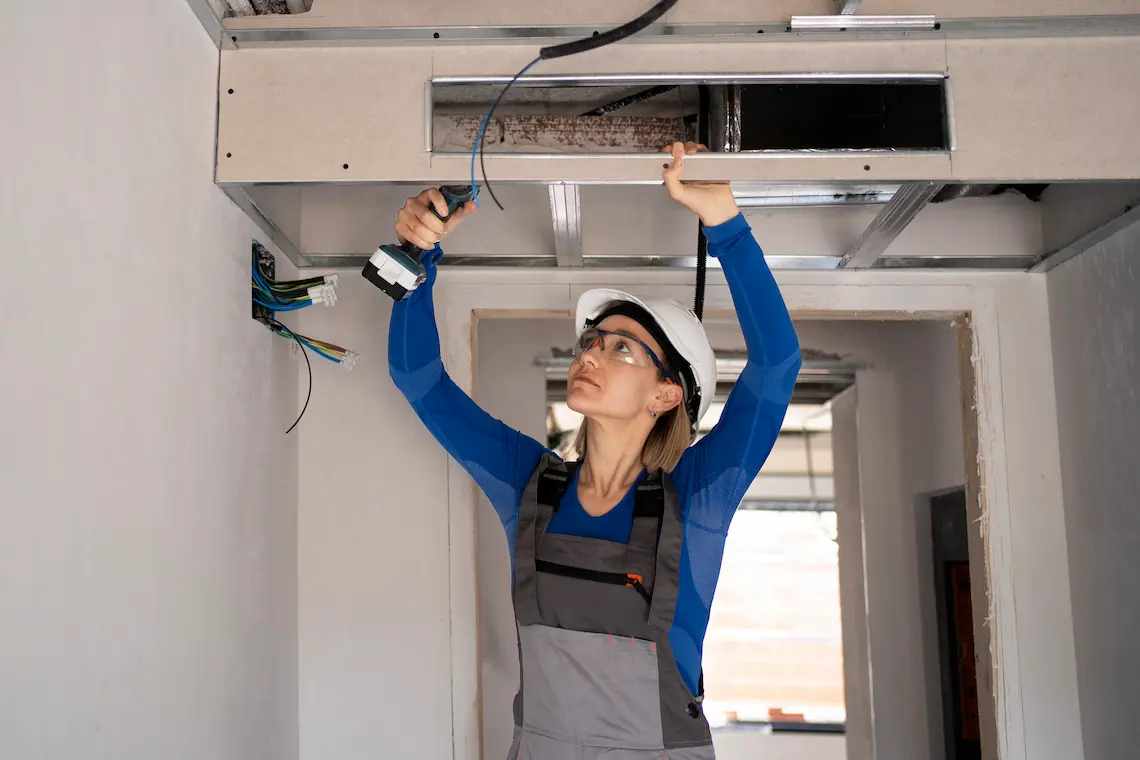 Female technician wearing safety gear using a power drill to fix indoor ductwork during a comprehensive ac repair brownsville tx service call.